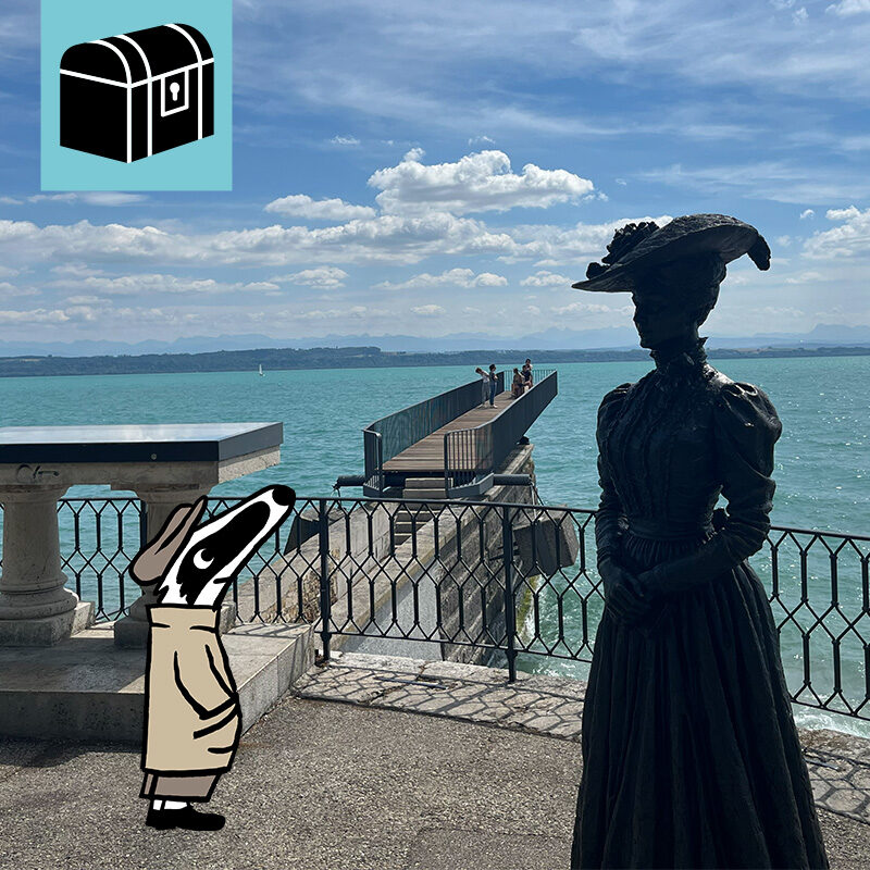 Auf dem Bild sieht man die Utopie Passarelle am Neuenburgersee. Im Vordergrund ist eine der beiden Figuren aus der Belle Époque zu sehen. Ausserdem wurde Detektiv Dachs dem Foto hinzugefügt, der die Figur ansieht.